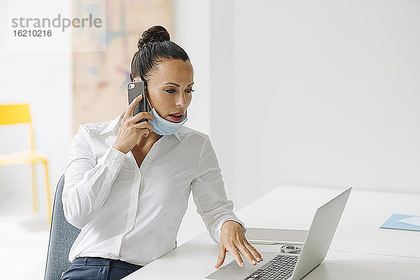 Geschäftsfrau mit Maske  die über ein Smartphone spricht  während sie einen Laptop auf dem Schreibtisch im Heimbüro benutzt