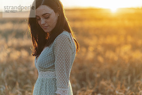 Nachdenkliche junge Frau  die bei Sonnenuntergang auf einem Bauernhof steht und nach unten schaut
