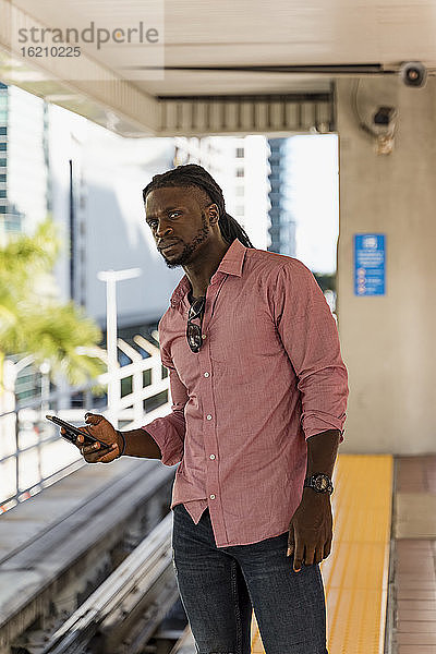 Junger Afroamerikaner  der sein Smartphone hält und wegschaut  während er am Bahnhof steht  Miami  Florida  USA
