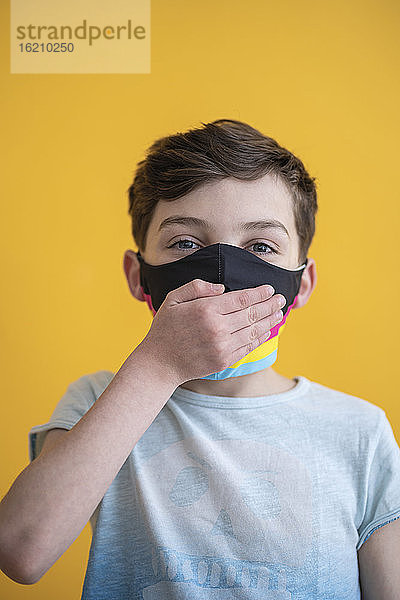 Nahaufnahme eines Jungen mit Maske  der den Mund mit der Hand bedeckt  vor gelbem Hintergrund