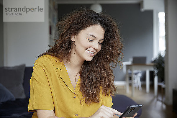 Junge Frau lächelt  während sie zu Hause eine Textnachricht auf ihrem Smartphone schreibt