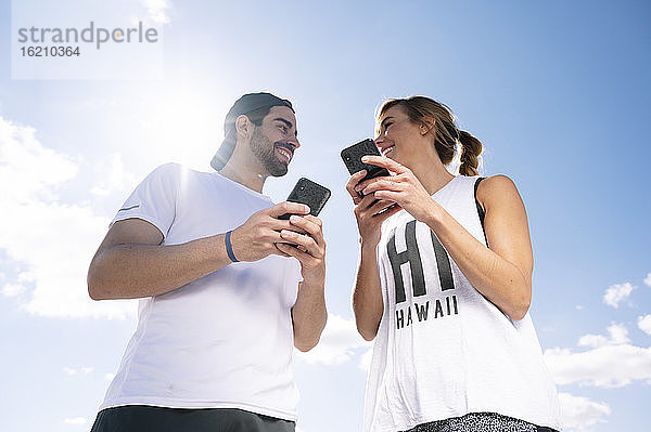 Lächelndes Paar  das Smartphones hält und sich gegenseitig anschaut  während es an einem sonnigen Tag vor dem Himmel steht