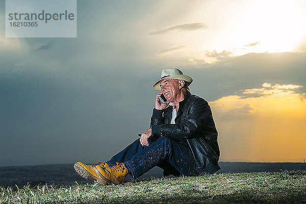 Lächelnder älterer Mann  der mit seinem Handy gegen den bewölkten Himmel bei Sonnenuntergang spricht