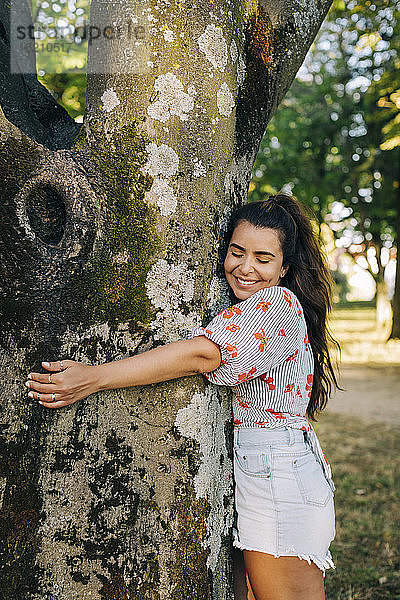 Lächelnde junge Frau mit geschlossenen Augen  die einen Baumstamm umarmt  während sie im Park steht