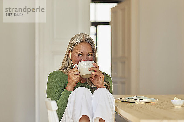 Lächelnde Frau trinkt Kaffee am Tisch zu Hause
