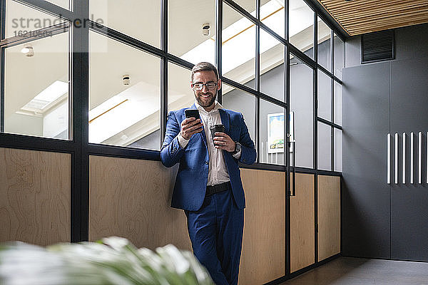 Lächelnder Geschäftsmann  der einen Einweg-Kaffeebecher hält  während er sein Smartphone an der Glaswand eines Coworking-Büros benutzt