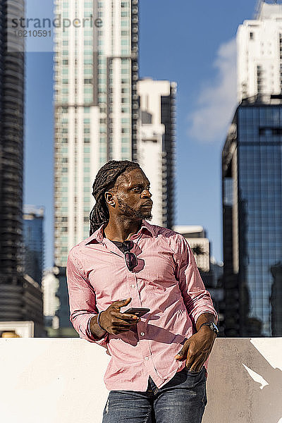 Junger Afroamerikaner  der sein Smartphone in der Hand hält und wegschaut  während er vor Wolkenkratzern in Miami  Florida  USA steht