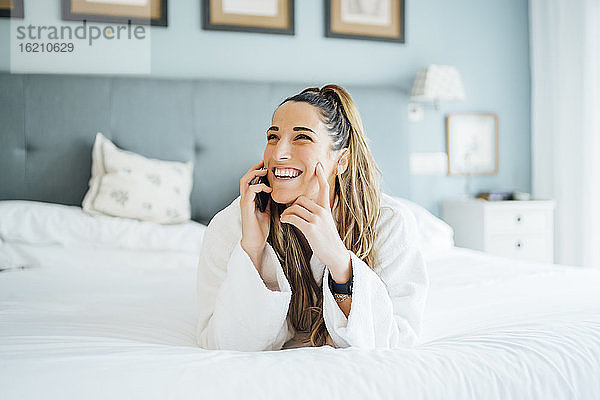 Fröhliche Frau im mittleren Erwachsenenalter  die sich über ein Smartphone unterhält  während sie auf einem Bett im Hotel liegt