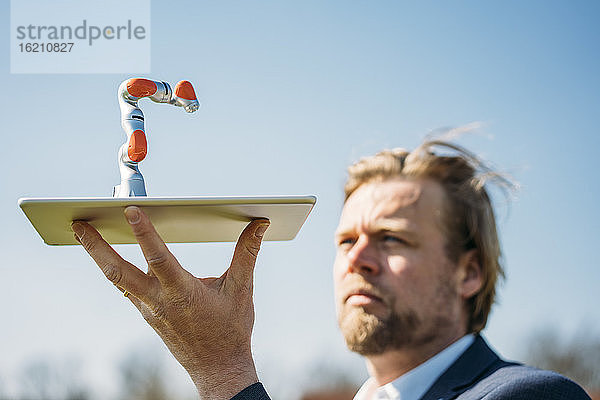 Männlicher Fachmann  der einen kleinen Roboterarm auf einem digitalen Tablet gegen den klaren Himmel hält