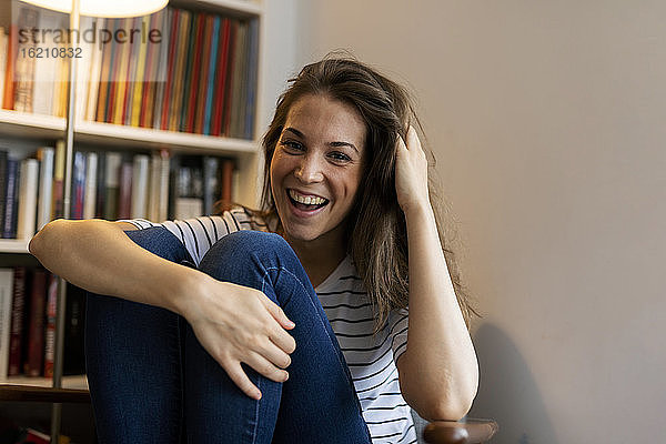 Fröhliche junge Frau mit Hand im Haar  die zu Hause an der Wand sitzt