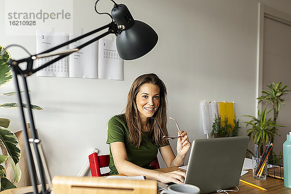 Lächelnde Geschäftsfrau mit Brille am Schreibtisch sitzend im Heimbüro
