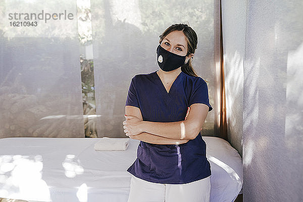 Weibliche Therapeutin mit Maske und verschränkten Armen steht am Massagetisch im Spa