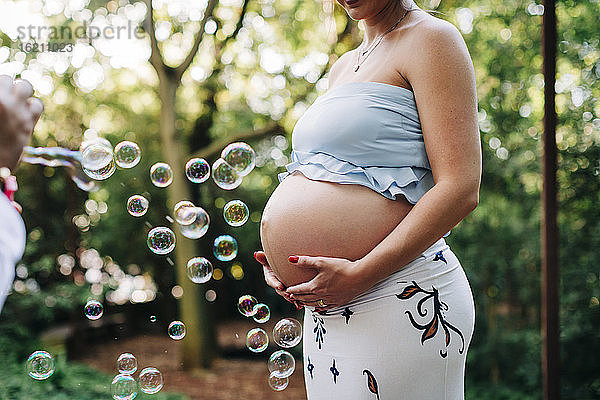 Schwangere Frau mit Händen auf dem Bauch steht bei Blasen im öffentlichen Park