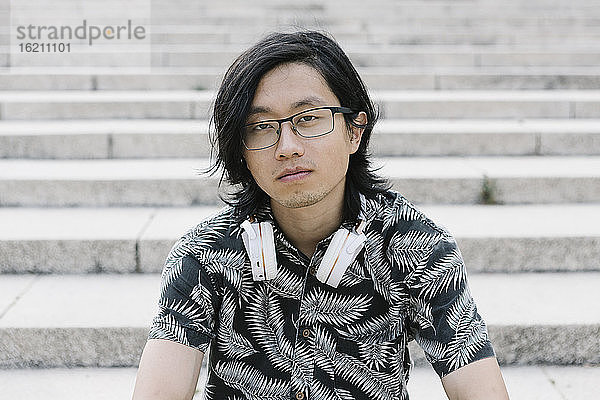 Nahaufnahme eines jungen Mannes mit Brille und Kopfhörern  der auf einer Treppe in der Stadt sitzt
