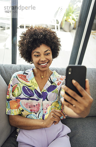 Glückliche Frau  die ein Selfie mit ihrem Smartphone macht  während sie auf dem Sofa im Penthouse sitzt