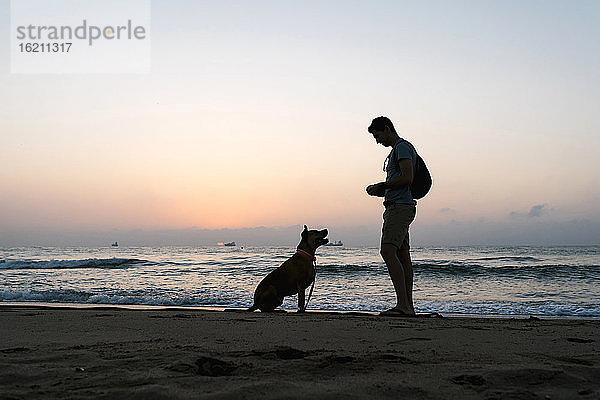 Silhouette Mann stehend mit seinem Hund am Strand