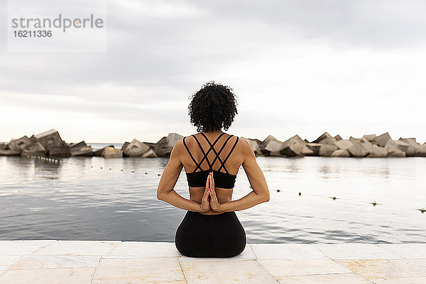 Mittlere erwachsene Frau mit lockigem Haar übt Yoga  während sie auf der Promenade am Meer sitzt