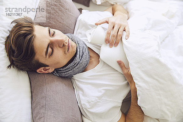 Nahaufnahme eines kranken jungen Mannes mit Wärmflasche  der zu Hause auf dem Bett schläft