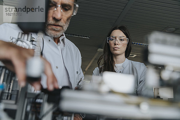 Männliche und weibliche Wissenschaftler mit Schutzbrillen bei der Untersuchung einer Maschine im Labor