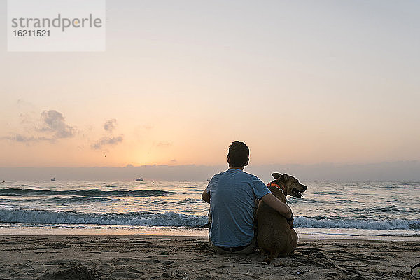 Mann sitzt mit seinem Hund und genießt die Morgendämmerung am Strand