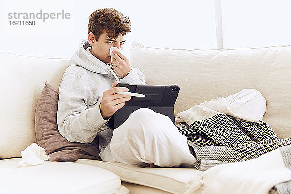 Junger Mann schnäuzt sich die Nase  während er ein digitales Tablet auf dem Sofa zu Hause benutzt