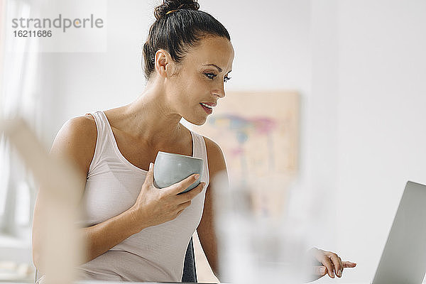 Frau hält Kaffeetasse und benutzt Laptop  während sie zu Hause im Büro an der Wand sitzt