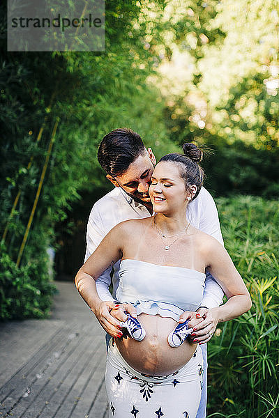 Romantisches schwangeres Paar  das sich im Park umarmt und Babyschuhe hält