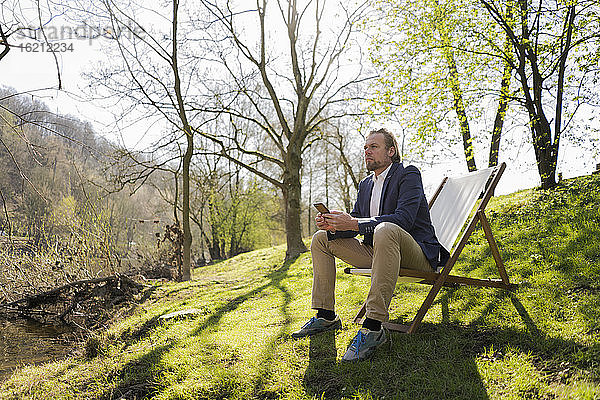 Nachdenklicher Geschäftsmann  der sein Smartphone in der Hand hält  während er auf einem Stuhl vor den Bäumen im Park sitzt