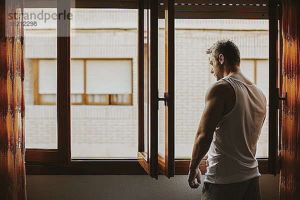 Nachdenklicher Mann im mittleren Erwachsenenalter  der durch ein Fenster schaut  während er zu Hause steht