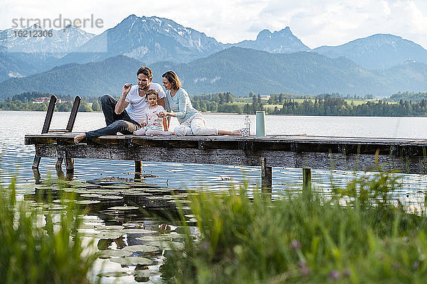 Eltern mit Tochter sitzen auf dem Steg über dem See vor den Bergen