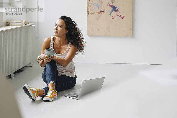 Nachdenkliche Frau  die eine Kaffeetasse hält  während sie mit ihrem Laptop zu Hause auf dem Boden sitzt