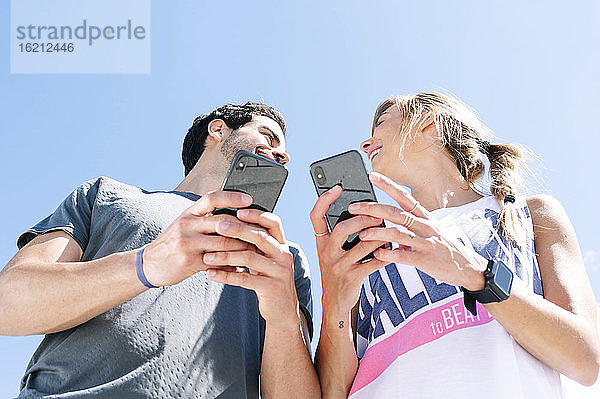 Lächelndes Paar  das sich gegenseitig ansieht  während es ein Smartphone gegen den klaren blauen Himmel benutzt