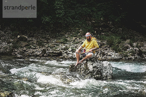 Abenteurer mit Bart sitzt in der Mitte des Flusses auf einem Stein und schnitzt an einem Stück Holz