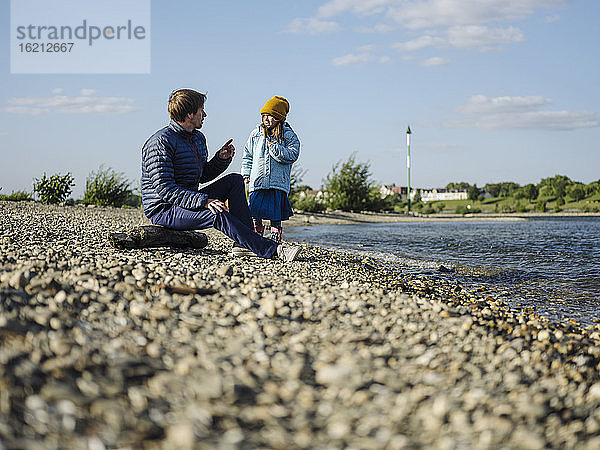 Vater spricht mit Tochter am Flussufer sitzend gegen den Himmel an einem sonnigen Tag