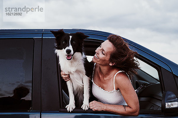 Lächelnde Frau  die einen Hund ansieht  während sie sich durch ein Autofenster lehnt