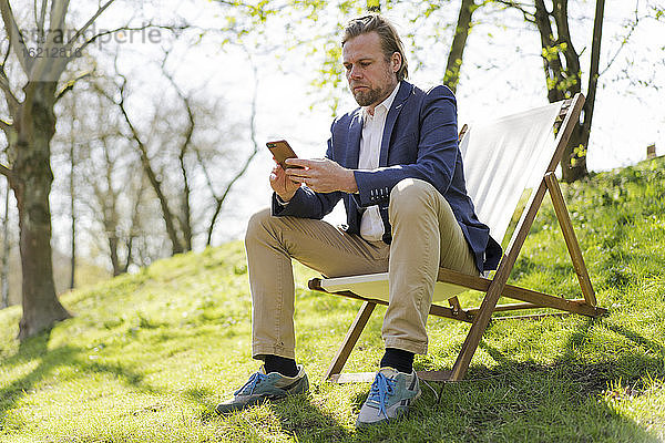 Männlicher Unternehmer  der sein Smartphone benutzt  während er auf einem Stuhl im Park sitzt