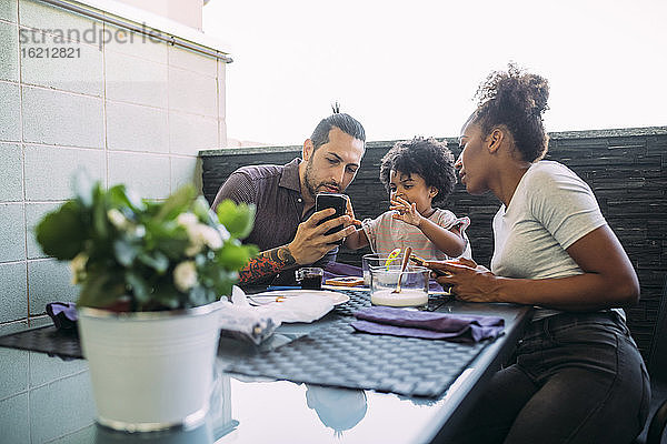 Mann zeigt Tochter und Frau am Esstisch sein Smartphone