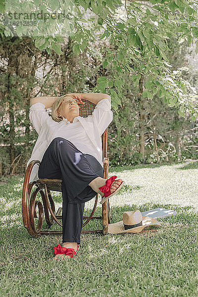 Nachdenkliche ältere Frau mit Händen hinter dem Kopf entspannt auf einem Stuhl im Hof