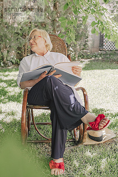 Ältere Frau hält ein Buch und schaut weg  während sie sich auf einem Stuhl im Garten entspannt
