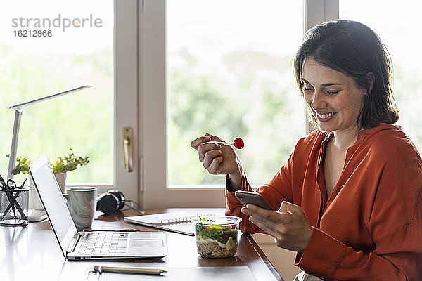 Lächelnder Geschäftsmann  der sein Mobiltelefon benutzt  während er zu Hause Salat isst