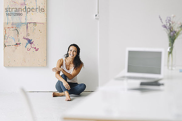 Fröhliche Frau im mittleren Erwachsenenalter  die zu Hause auf dem Boden an der Wand im Loft sitzt