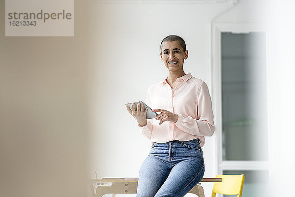 Porträt einer lächelnden Geschäftsfrau  die in einem Loftbüro auf einem Schreibtisch sitzt und ein Tablet hält