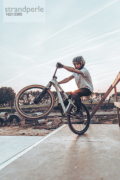 Junger Mann mit Helm fährt Fahrrad auf einer Rampe im Park gegen den Himmel bei Sonnenuntergang