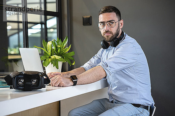 Gut aussehender Geschäftsmann sitzt mit Headset und Laptop am Schreibtisch in einem kreativen Coworking-Büro