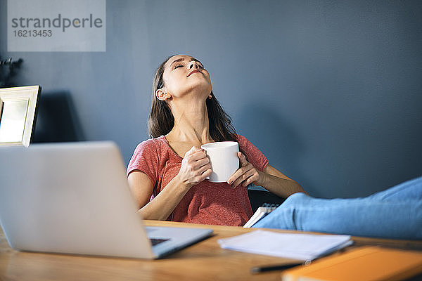 Unternehmerin hält Kaffeetasse  während sie sich am Schreibtisch im Heimbüro entspannt