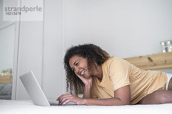 Fröhliche Frau mit lockigem Haar  die einen Laptop benutzt  während sie zu Hause auf dem Bett liegt