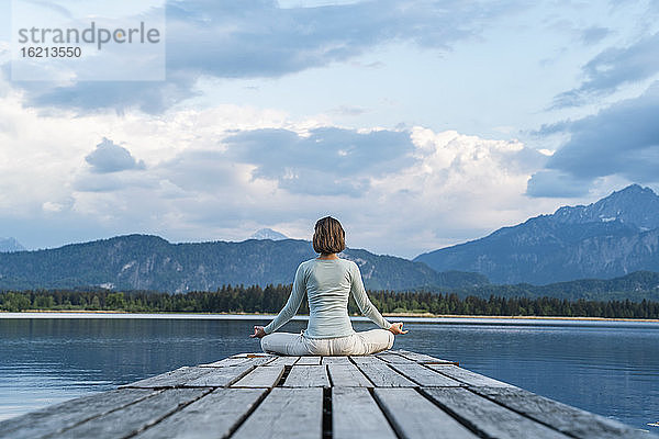 Mittlere erwachsene Frau meditiert sitzend auf einem Steg über einem See vor bewölktem Himmel