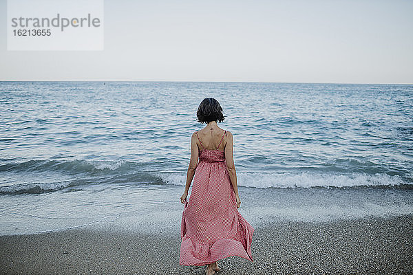 Frau im Kleid geht am Strand gegen den klaren Himmel bei Sonnenuntergang zum Meer