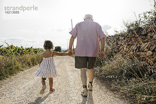 Großvater hält die Hand seiner Enkelin beim Spaziergang auf einem unbefestigten Weg gegen den Himmel
