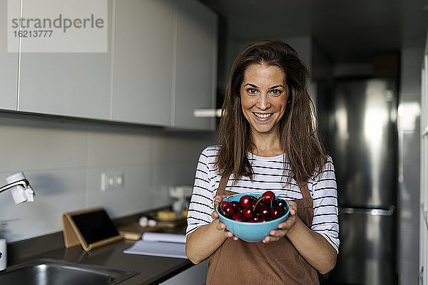 Lächelnde junge Frau  die Kirschen in der Hand hält  während sie in der Küche zu Hause steht
